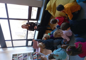 Dzieci siedzą na dywanie w bibliotece i odpowiadają na pytania pani bibliotekarki